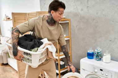 Dövmeleri olan yakışıklı bir genç adam evde çamaşır yıkamaya hazırlanan bir çamaşır sepeti taşıyor..