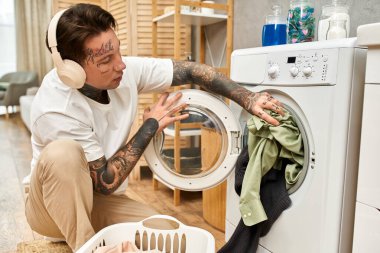 Genç, dövmeli bir adam rahat bir evde çamaşır makinesine çamaşır yüklüyor..