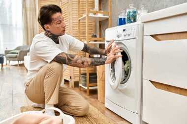 Dövmeli bir adam çamaşır makinesinin önünde diz çöker, sıcak, davetkar bir yerde çamaşırlarla meşgul olur..