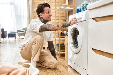 Günlük kıyafetli dövmeli adam evde çamaşır makinesi ayarlarını ayarlamaktan mutluluk duyar..