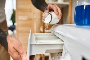 Yakışıklı dövmeli bir adam çamaşır makinesinin çekmecesine deterjan döker. Ev rutininin keyfini çıkarır..