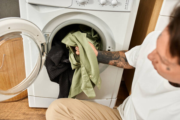 Tattooed man sorts clothes into a washing machine, embracing comfort in his cozy home setting.