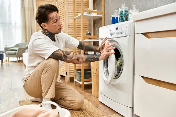 Dövmeli bir adam çamaşır makinesinin önünde diz çöker, sıcak, davetkar bir yerde çamaşırlarla meşgul olur..