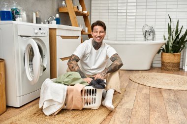 Sıcak bir evde, çekici dövmeli bir adam çamaşırları sıralarken sıcacık gülümser..