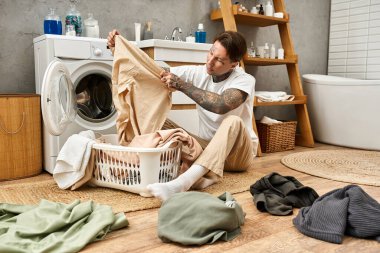 Dövmeleri olan genç bir adam rahat bir şekilde yerde oturuyor, rahat bir banyoda renkli çamaşırları sıralıyor..