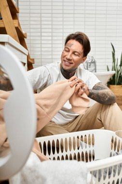 Dövmeleri olan genç bir adam evinde rahat bir şekilde oturur, çamaşırları neşeli bir ifadeyle katlar..