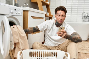 Dövmeleri olan genç bir adam yerde rahat bir şekilde oturur, çamaşırları yıkarken mesajlaşır..