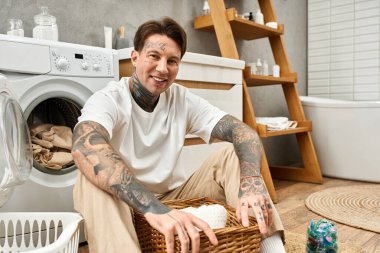 Rahatlamış dövmeli bir adam yerde oturur, sıcak ve davetkar bir evde çamaşırları sıralar..