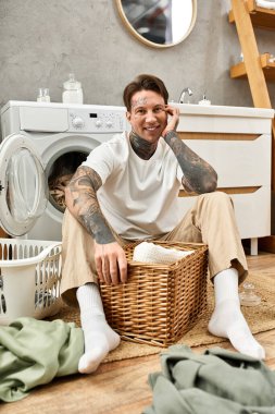 Dövmeli yakışıklı genç adam evde rahatlıyor, çamaşır sepetinin yanında oturuyor, sıcak bir şekilde gülümsüyor..