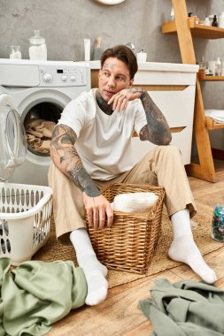 Dövmeleri olan genç bir adam evde rahatlıyor, sıcak bir atmosferde çamaşırlarını sıralıyor..