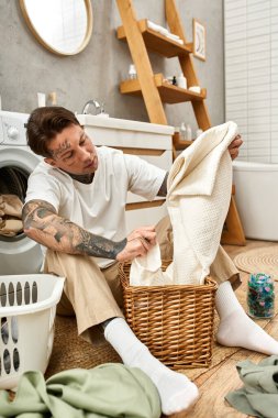 Dövmeli genç bir adam, rahat evinde çamaşırların arasında havluyu düzgünce katlıyor..