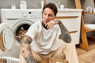 Rahatlamış dövmeli adam çamaşır makinesinin yanında rahatça oturur, evde rahat bir anın tadını çıkarır..