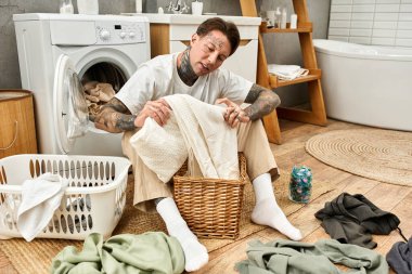 Genç, dövmeli bir adam çamaşırları evde dizer, sıcaklık ve rahatlama yayar..