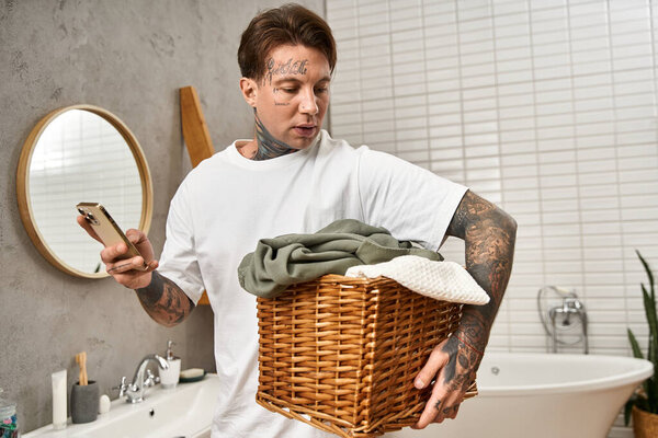 Tattooed man smiles with laundry basket, checking his phone in a cozy home setting.