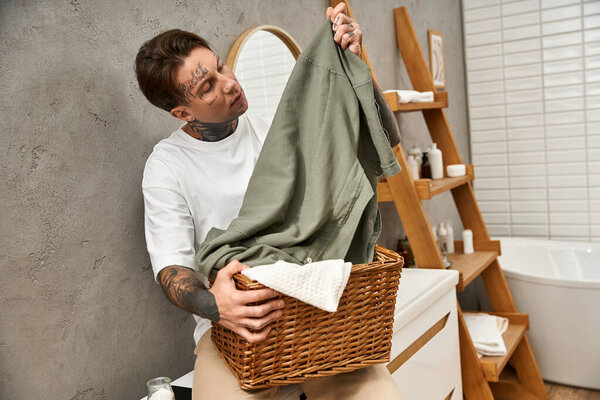 A young man with tattoos meticulously organizes laundry in a rustic basket at home.