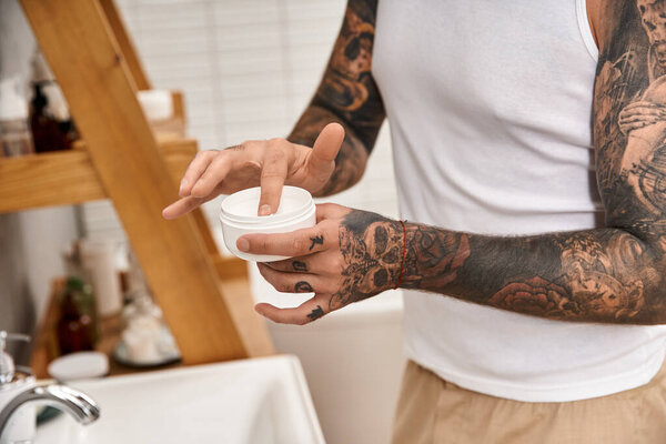 A young tattooed man enjoys his time at home, applying cream during a relaxing self care routine.
