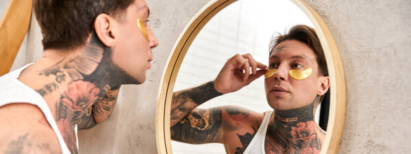 A young man puts on under eye patches, gazing at his reflection and feeling relaxed.