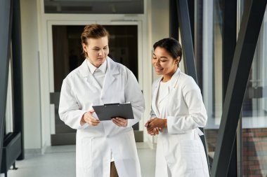 Two female physicians engaged in a discussion while walking through a contemporary hospital.