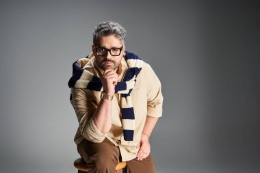 Keskin gözlüklü yakışıklı bir adam zarif bir stüdyoda düşünceli bir duruşu olan modaya uygun bir kıyafet modelliyor..