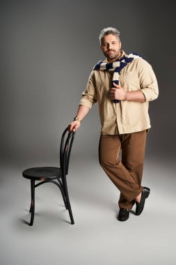Tüylü şık bir adam kendine güvenerek bir sandalyenin yanında duruyor, şık kıyafetiyle hava atıyor..