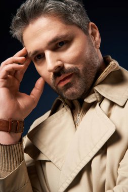 Kendine güvenen bir adam modaya uygun bir trençkotla poz veriyor, stüdyo ortamında çağdaş tarzın altını çiziyor..