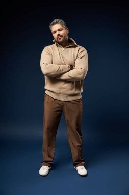 Tüylü, kendine güveni olan bir adam, şık bir kıyafetle karanlık bir stüdyo arkaplanına karşı poz verir..