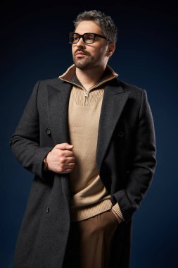 Gözlüklü yakışıklı bir adam şık kıyafetlerini şık bir stüdyo ortamında sergiliyor..