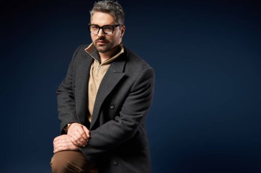 Şık bir adam, sakin bir stüdyo ortamında gözlüklü, modern ve zarif bir kıyafet sergiliyor..