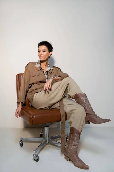 Full length of archaeologist in safari style clothes and cowboy boots sitting on office chair and looking away on grey — Stock Photo