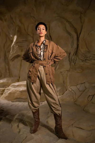 Full length of brunette archaeologist in brown jacket and pants standing with hands on hips in cave — Stock Photo