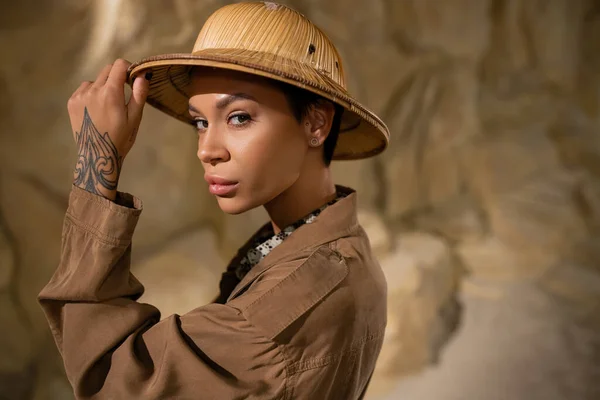 Arqueólogo tatuado en chaqueta marrón ajustando sombrero de safari y mirando a la cámara en el desierto - foto de stock