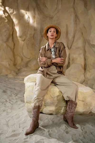 Joven arqueólogo en traje de safari sentado en piedra con los brazos cruzados cerca de la roca - foto de stock