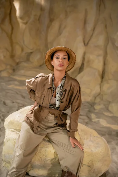 Archäologe im Safari-Outfit sitzt mit der Hand auf der Hüfte auf einem Felsen und blickt in die Kamera — Stockfoto
