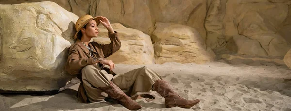 Longitud completa de joven arqueólogo sentado en la arena cerca de piedra y el ajuste de sombrero de safari, bandera - foto de stock
