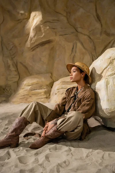 Archaeologist in safari hat and beige clothes with cowboy boots sitting near stone in cave — Stock Photo