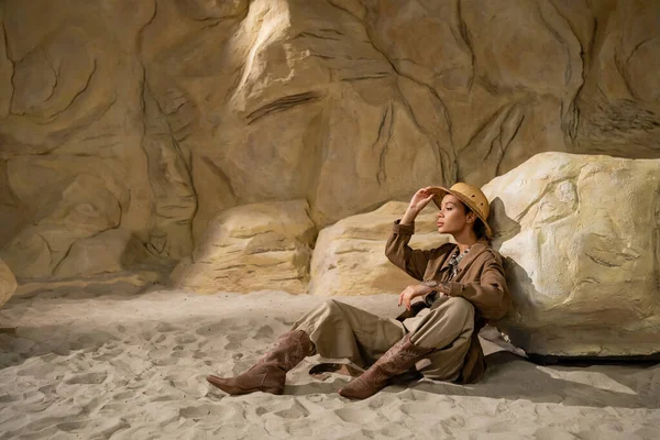 Comprimento total do arqueólogo ajustando chapéu de safári enquanto sentado na areia perto de pedra — Fotografia de Stock