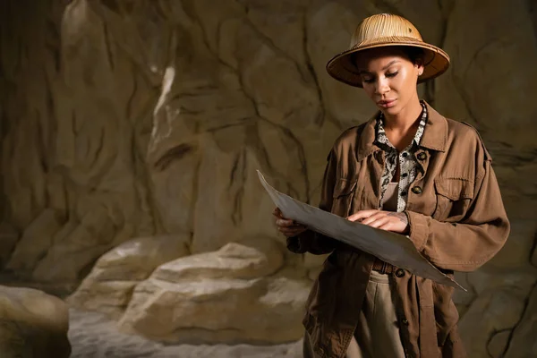 Archäologe in brauner Jacke und Safarimütze betrachtet alte Landkarte in Höhle — Stockfoto