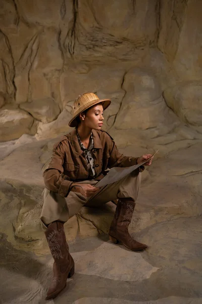 Археолог в сафарі капелюсі і ковбойських черевиках, дивлячись на стародавню карту в печері — стокове фото