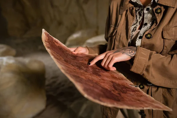 Vista recortada de arqueólogo en chaqueta marrón apuntando al mapa antiguo en la cueva - foto de stock