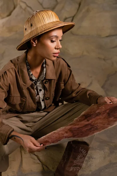 Archaeologist in safari style outfit looking at ancient map while sitting in cave — Stock Photo
