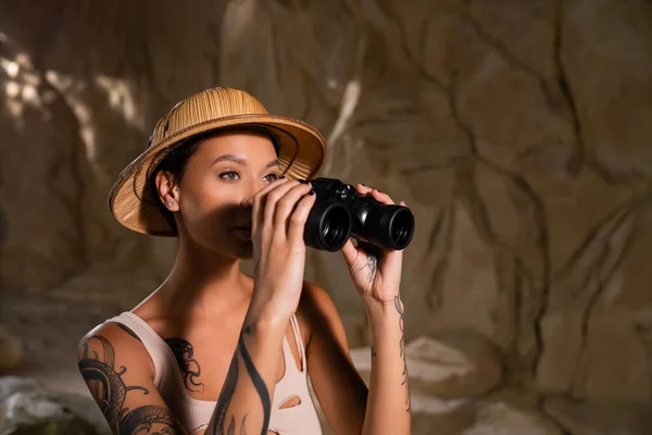 Tattooed explorer in safari hat looking in binoculars during archaeological expedition — Stock Photo