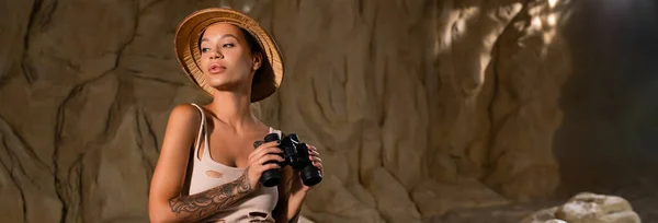 Joven arqueólogo en top sexy y sombrero de safari sosteniendo binoculares en el desierto, pancarta - foto de stock