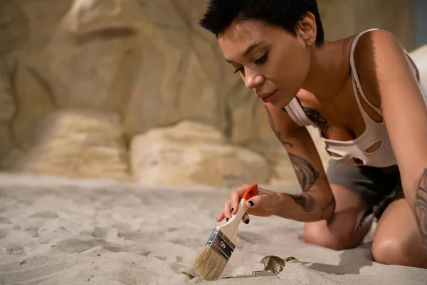 Bastante morena arqueólogo sosteniendo cepillo cerca de artefacto en arena - foto de stock