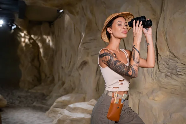 Sexy and tattooed archaeologist in safari hat holding binoculars and looking at camera in desert — стоковое фото
