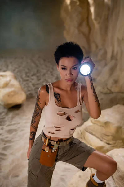 Tattooed brunette archaeologist with waist bag holding flashlight in cave — Stock Photo