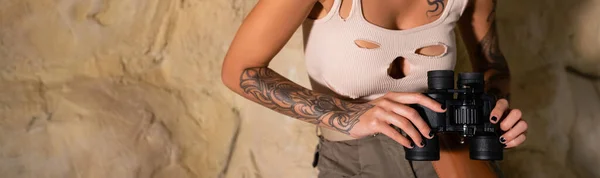 Vista parcial del arqueólogo tatuado en la parte superior del tanque sexy sosteniendo binoculares en el desierto, bandera - foto de stock
