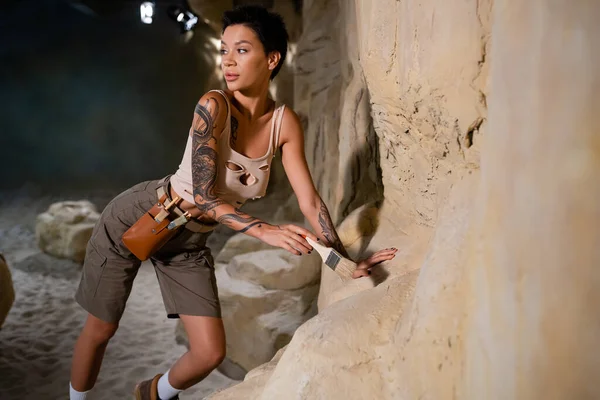 Sexy arqueólogo tatuado sosteniendo cepillo cerca de la roca y mirando hacia otro lado - foto de stock