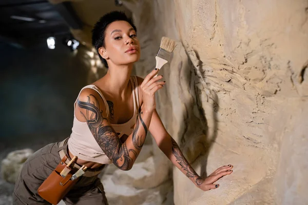 Tätowierte und sexy Archäologin blickt in die Kamera, während sie den Pinsel in der Nähe von Felsen in der Höhle hält — Stockfoto