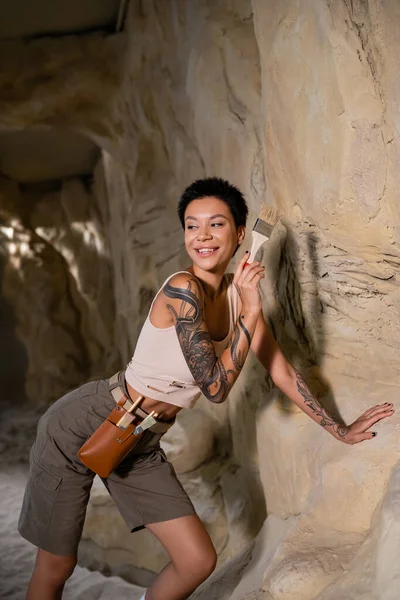 Sonriente arqueólogo tatuado en camiseta sin mangas mirando hacia otro lado mientras trabaja con cepillo cerca de roca en cueva - foto de stock