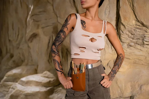 Partial view of tattooed archaeologist in tank top and waist bag with brushes standing with hands on hips near rock — Stock Photo